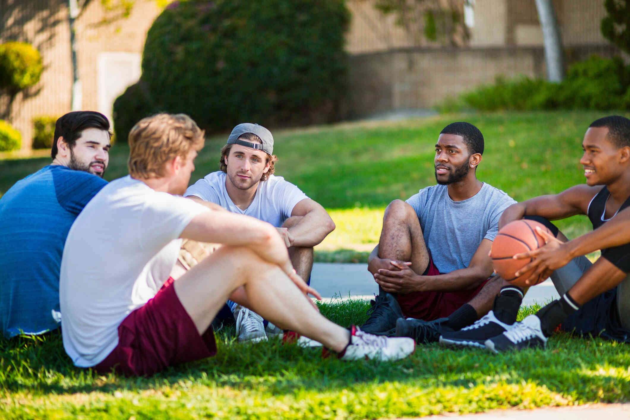 A group of men are seated on the ground in a circle seems chatting casually, while one person holding a basketball.